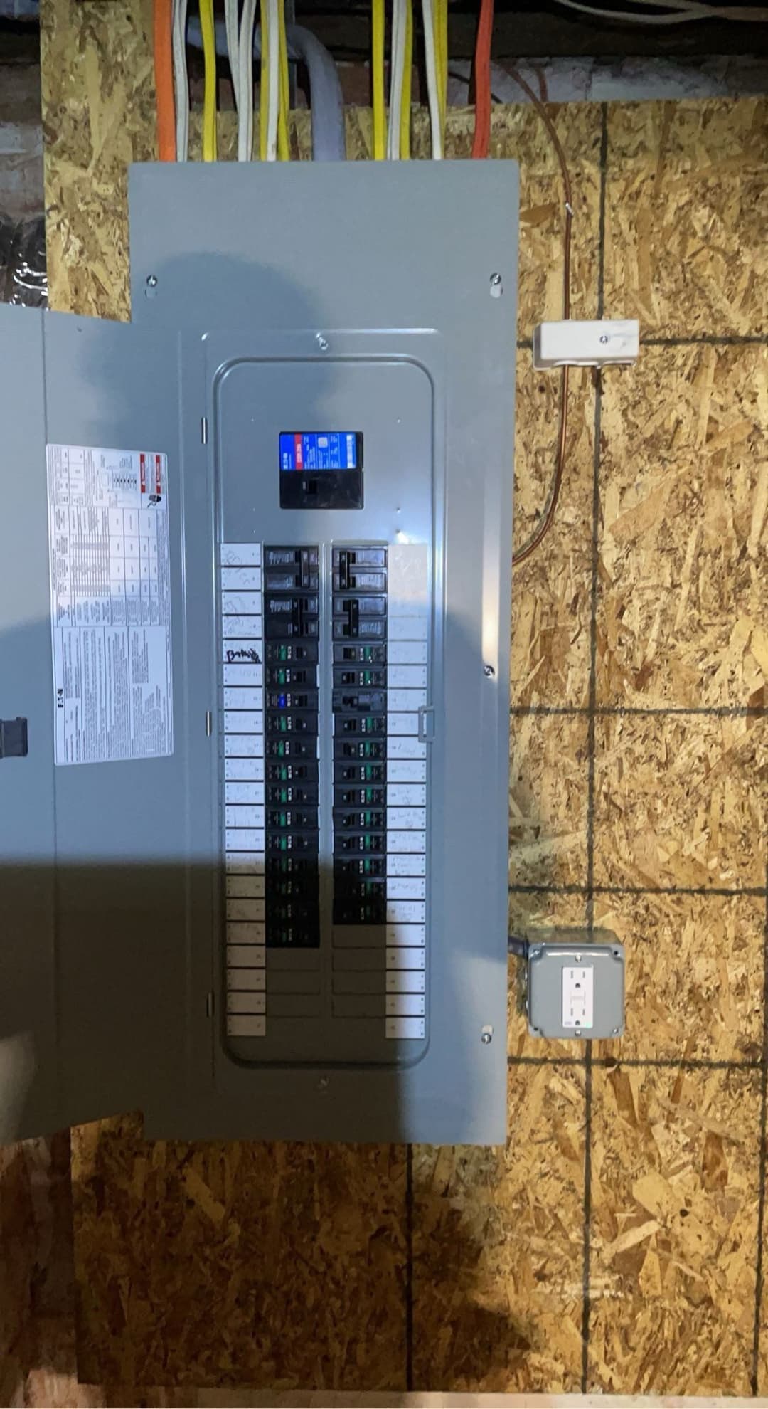 Electrical breaker panel with circuit labels and digital display mounted on wooden wall.