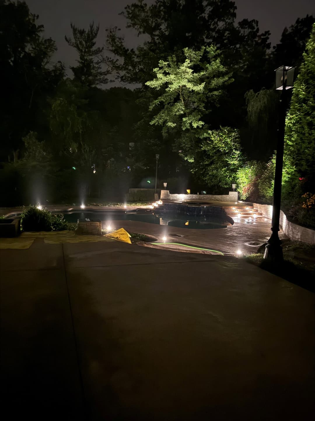 Nighttime view of a beautifully lit backyard pool area surrounded by lush greenery.