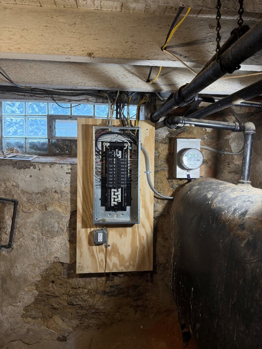 Electrical panel mounted on wooden board in a basement with pipes and glass block window.