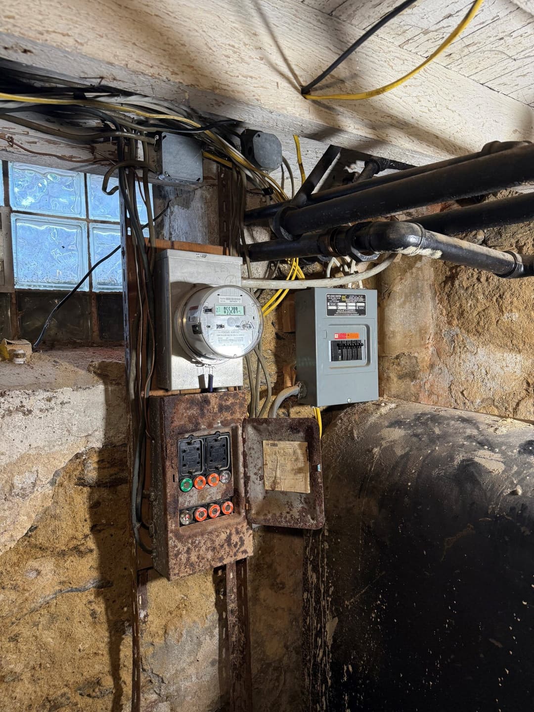 Electrical meter and circuit panel in a rustic basement with exposed wiring.