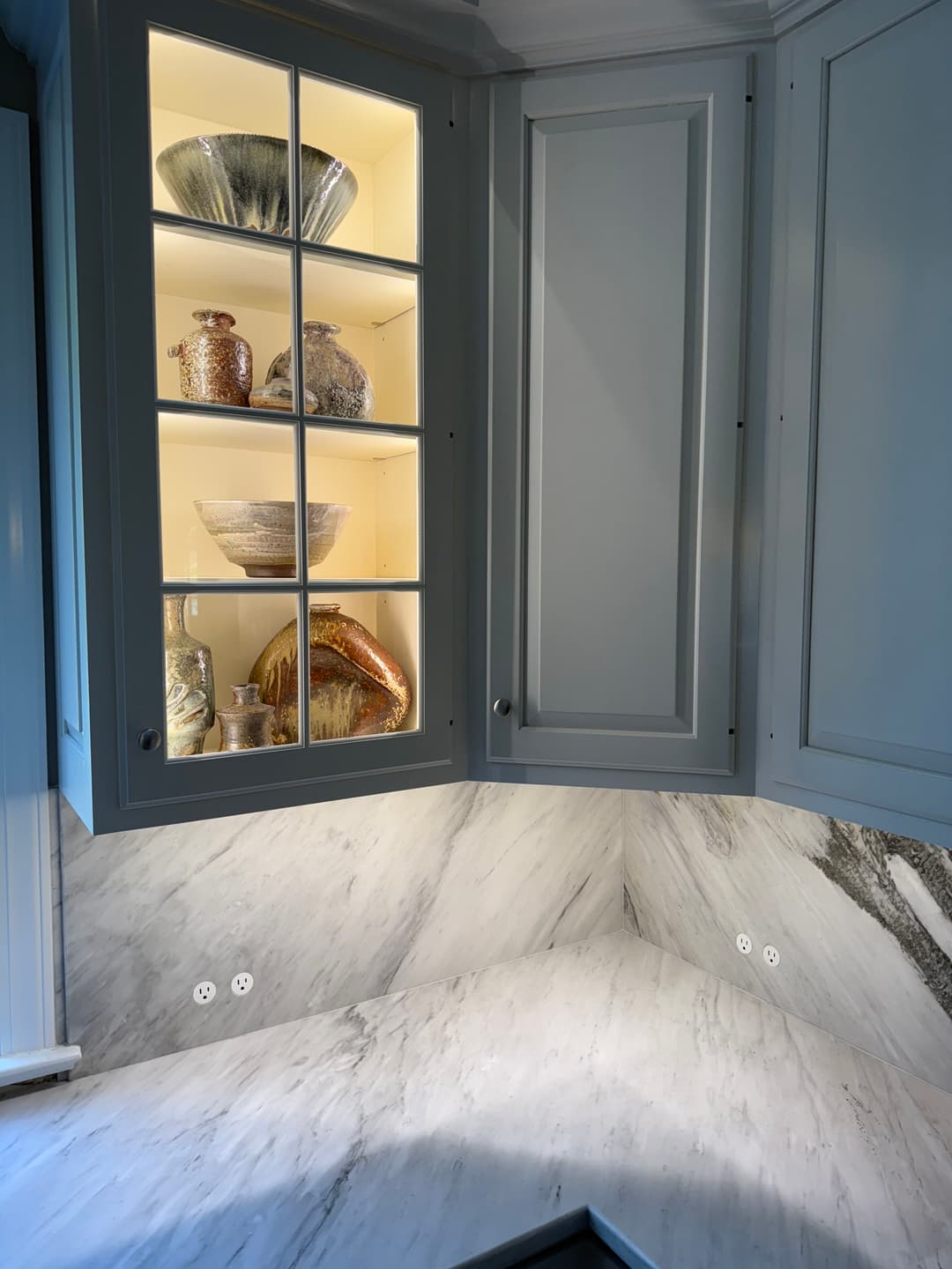 Modern kitchen corner cabinet with illuminated glass shelves displaying decorative glassware.