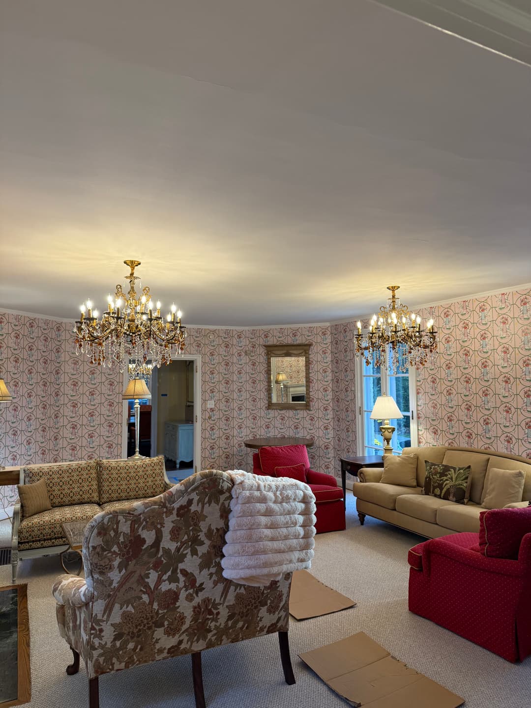 Living room with floral chairs, red sofas, chandeliers, and patterned wallpaper.